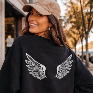 May include: Black crewneck sweatshirt with a white wing design on the back. The sweatshirt is worn with denim shorts and a tan baseball cap. The image is taken outdoors on a sunny day.