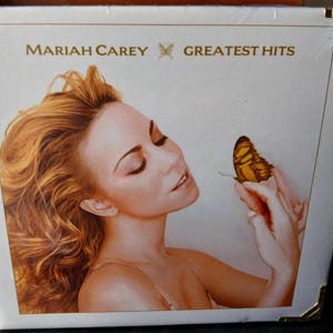 Könnte beinhalten: Das Cover des Albums Greatest Hits von Mariah Carey zeigt ein Porträt der Sängerin mit rötlichem Haar, die einen Schmetterling hält. Der Albumtitel und der Name des Künstlers sind in Gold auf weißem Hintergrund gedruckt. Das Album ist in Plastik versiegelt.