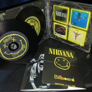 Könnte beinhalten: Ein Nirvana CD-Set mit Albumcover und Bandlogo. Das Set enthält CDs, ein Booklet mit Bandfotos und eine transparente Kunststoffhülle. Das Albumcover zeigt ein gelbes Smiley-Gesicht und den Namen der Band.