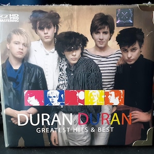 Peut inclure: Pochette d'album du groupe Duran Duran. L'image présente les cinq membres du groupe. Le titre de l'album "Greatest Hits & Best" est affiché avec le nom du groupe en lettres colorées.