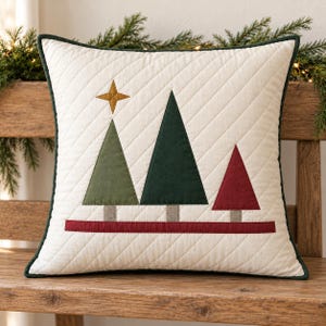 May include: A square throw pillow with a cream-colored quilted cover featuring diagonal stitching. The design includes three triangular trees in olive green, dark green, and red, with a gold star above and a horizontal red stripe.