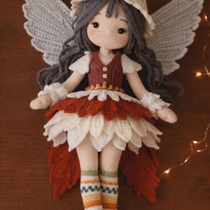 Puede incluir: Muñeca de hada de ganchillo hecha a mano, con cabello gris, un sombrero a rayas y grandes alas grises. La muñeca lleva una falda de pétalos roja y blanca, un chaleco burdeos y calcetines a rayas con zapatos rojos. Un artículo artesanal caprichoso.