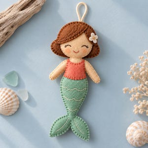 Puede incluir: Un adorno de fieltro con forma de sirena que presenta cabello castaño con una pequeña flor blanca, un top color coral y una cola verde claro con detalles de escamas cosidas. La figura tiene una cara sonriente y un lazo blanco para colgar.