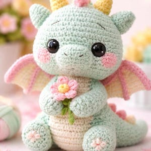 Puede incluir: Un peluche de dragón tejido a ganchillo en colores pastel suaves. El dragón es principalmente verde menta con vientre y detalles crema. Tiene grandes ojos negros, mejillas rosadas y sostiene una flor rosa. Las alas son rosas y amarillas.