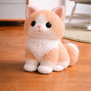 Puede incluir: Figura de gato de ganchillo hecha a mano en tonos melocotón y blanco. El gato tiene ojos negros grandes, nariz rosa y pecho blanco y esponjoso. El gato está sentado sobre una superficie de madera.