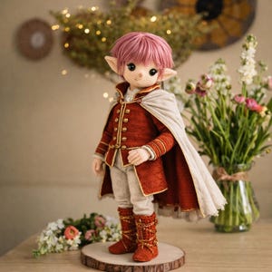 Puede incluir: Muñeca de ganchillo hecha a mano que representa a un elfo. La muñeca viste una chaqueta roja con detalles dorados, una capa crema y botas rojas. Tiene pelo rosa y orejas puntiagudas. La muñeca está sobre una base de madera.