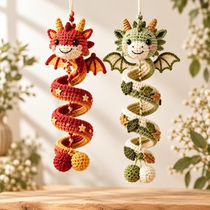 Puede incluir: Dos adornos colgantes de dragón hechos a ganchillo, uno en rojo y amarillo con estrellas, y otro en verde y crema con hojas. Ambos tienen cuerpos en espiral, pequeñas alas, cuernos y cabezas redondas con ojos negros y mejillas rosadas.