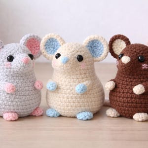 Puede incluir: Tres ratones de ganchillo hechos a mano en gris, crema y marrón. Cada ratón tiene ojos negros grandes y redondos, detalles rosas o azules en las orejas y patas, y una cola larga. Los ratones se exhiben sobre una superficie de madera clara.