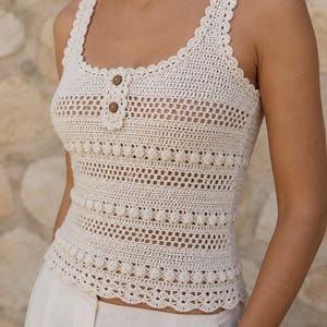 Peut inclure: Un haut sans manches en crochet de couleur crème doté d'une encolure dégagée avec un bord festonné, deux petits boutons marron sur la poitrine, et des rangées horizontales de points bobble texturés alternant avec des motifs ajourés.