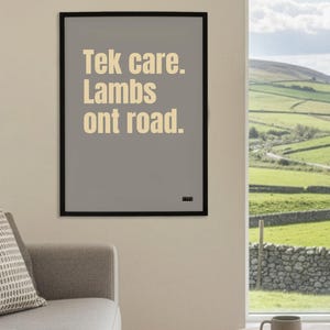Puede incluir: Un póster enmarcado con el texto "Tek care. Lambs ont road." en beige claro sobre un fondo gris. El póster está en un marco negro y está colgado en una pared. La habitación tiene una ventana con vistas a colinas verdes.