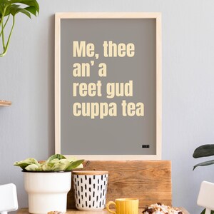 Puede incluir: Un cuadro enmarcado con el texto "Me, thee an' a reet gud cuppa tea" en una fuente beige claro sobre un fondo gris. El marco es de madera clara. El cuadro se muestra con plantas y una taza amarilla.