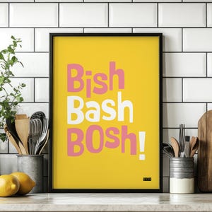 Puede incluir: Un cuadro enmarcado con las palabras "Bish Bash Bosh!" en rosa y blanco sobre un fondo amarillo. El cuadro está en un marco negro y se exhibe en un entorno de cocina con utensilios y limones.