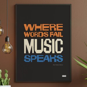 Puede incluir: Póster enmarcado en negro con el texto "WHERE WORDS FAIL MUSIC SPEAKS" en naranja, crema y azul. El póster está colgado en una pared marrón, con dos bombillas colgantes y una planta en maceta en primer plano. El póster incluye el texto "Hans Christian Andersen" y la palabra "TYPE".
