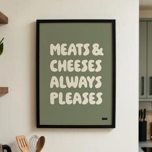 May include: A rectangular wall art print in a black frame featuring a sage green background with the off-white text MEATS & CHEESES ALWAYS PLEASES in a bold, wavy, retro-inspired font. The design is centred within the frame.