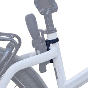 May include: A black bicycle lock attached to the white frame of a bicycle. The lock is secured with a black strap. A black bicycle rack is visible in the background, along with a portion of the rear tire.