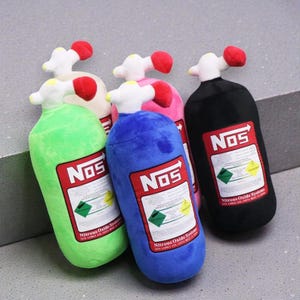 Puede incluir: Un conjunto de botellas de peluche de sistema de óxido nitroso (NOS) en varios colores, incluyendo verde, azul, rosa, beige y negro. Cada botella presenta una parte superior blanca con detalles rojos y una etiqueta roja y blanca con el logo "NOS" e información de seguridad.