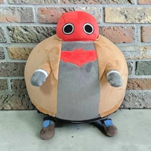 May include: A round plush toy featuring a red head with large black and white eyes, wearing a tan jacket over a grey torso with a red bat-shaped emblem. The figure has small grey arms, brown shoes with blue accents, and a spherical body shape.
