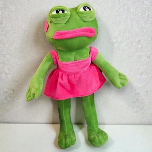 Könnte beinhalten: Eine grüne Plüschfroschpuppe mit einem rosa Kleid und großen rosa Lippen. Der Frosch hat einen skurrilen Ausdruck mit großen, schläfrigen Augen und einer kleinen rosa Blume auf der Wange. Die Puppe steht aufrecht.