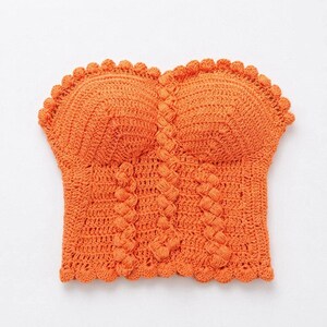 May include: An orange crochet top with a scalloped edge and braided detailing. The top features a sweetheart neckline and a fitted silhouette, perfect for summer wear. The handmade garment is a vibrant, eye-catching piece.