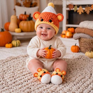 May include: A crochet baby hat and matching booties set featuring a candy corn design with white, orange, and yellow yarn, small round ears, and black button eyes. The hat has a pom-pom on top, and the booties mirror the same color scheme.