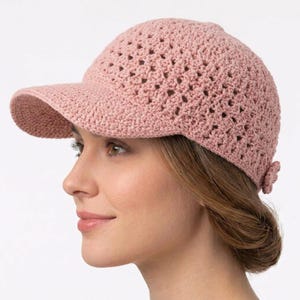 May include: A dusty pink baseball cap crafted from crocheted yarn featuring an openwork diamond pattern across the crown and a solid, tightly stitched brim. The cap includes a small, decorative crocheted flower detail at the back adjustment strap.