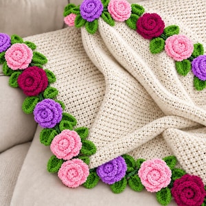 May include: A cream-colored crocheted blanket featuring a decorative border of three-dimensional flowers. The floral trim consists of pink, magenta, and purple crocheted roses interspersed with small green crocheted leaves along the edges.