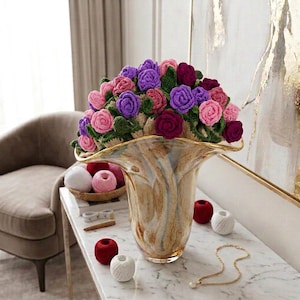 Puede incluir: Un jarrón decorativo con borde dorado, lleno de un ramo de rosas de ganchillo en rosa, morado y burdeos. El jarrón está sobre una mesa de mármol. También hay ovillos de lana y un collar sobre la mesa.