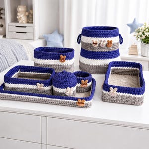 Puede incluir: Un conjunto de cestas de almacenamiento de ganchillo hechas a mano en azul, blanco y gris. El conjunto incluye una cesta grande con asas, varias cestas más pequeñas y una bandeja. Cada pieza está adornada con un acento decorativo de conejito de madera.