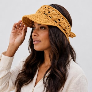 May include: A mustard yellow crochet visor featuring a detailed floral pattern across the headband and a solid brim. The accessory is tied at the back with matching yarn strings, creating a textured look with intricate stitching throughout the design.