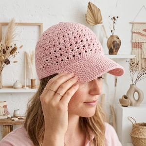 May include: A light pink baseball cap crafted from crocheted yarn featuring a textured, open-stitch pattern across the crown and a solid, tightly crocheted brim. The cap has a rounded shape and is constructed from a soft, matte-finish textile material.