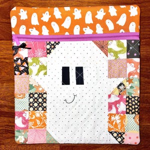 May include: A rectangular zippered pouch featuring a top band of orange fabric with white ghost patterns. The main body is a quilted white ghost shape with black eyes and a stitched smile, surrounded by a patchwork border of Halloween-themed prints.