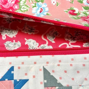 May include: A close-up of a quilted pouch with a pink zipper. The interior lining features a pink background with white kittens. The exterior has a floral pattern with pink and blue flowers. The bottom section has a patchwork design.