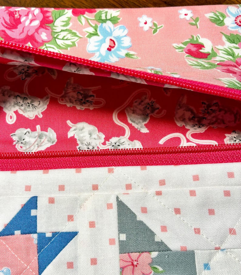 May include: A close-up of a quilted pouch with a pink zipper. The interior lining features a pink background with white kittens. The exterior has a floral pattern with pink and blue flowers. The bottom section has a patchwork design.