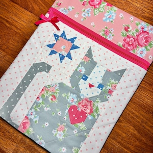 May include: A quilted fabric laptop sleeve featuring a cat design with a heart. The sleeve has a pink zipper and a pink bow. The cat is made of gray fabric with floral patterns, and the background is white with pink polka dots.