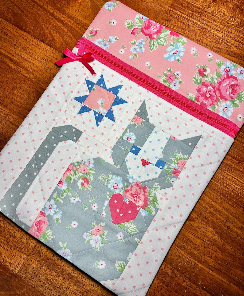 May include: A quilted fabric laptop sleeve featuring a cat design with a heart. The sleeve has a pink zipper and a pink bow. The cat is made of gray fabric with floral patterns, and the background is white with pink polka dots.