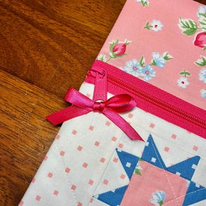 May include: Close-up of a quilted pouch with a pink floral pattern and a blue star design. The pouch features a pink zipper and a matching ribbon bow. The background is a wooden surface.