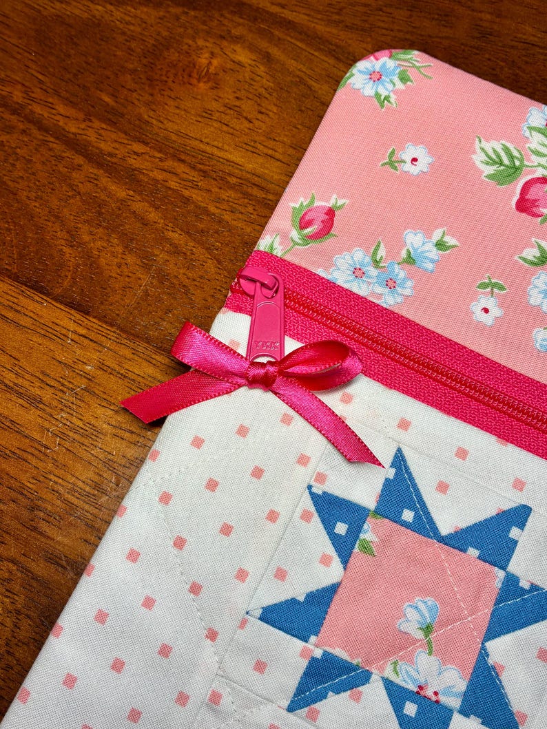 May include: Close-up of a quilted pouch with a pink floral pattern and a blue star design. The pouch features a pink zipper and a matching ribbon bow. The background is a wooden surface.