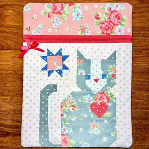 May include: A quilted fabric case with a pink floral top and a red zipper. The case features a patchwork cat design with floral patterns and a red heart. A blue star is also part of the design. The case is approximately 10 inches wide and 12 inches tall.