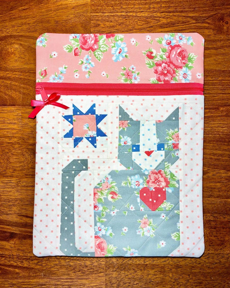 May include: A quilted fabric case with a pink floral top and a red zipper. The case features a patchwork cat design with floral patterns and a red heart. A blue star is also part of the design. The case is approximately 10 inches wide and 12 inches tall.
