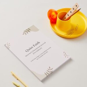 Könnte beinhalten: Ein Andachtstagebuch mit dem Titel "Quiet Faith" mit hellfarbigem Einband und floralen Akzenten. Ein gelber Schreibtisch-Organizer enthält einen Bleistift, ein Lineal und Büroklammern. Eine kleine orangefarbene Dekoration befindet sich in der Nähe.