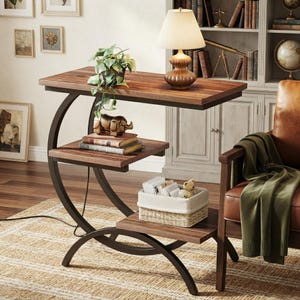 May include: A C-shaped side table with a dark metal frame and three wooden shelves. The top shelf holds a lamp and a potted plant, the middle shelf holds a gold-coloured rhinoceros figurine and two books, and the bottom shelf holds a woven basket.