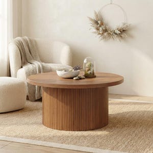 May include: A round wooden coffee table with a cylindrical base featuring vertical slats. The tabletop is smooth and brown, supporting a speckled white bowl containing dried lavender and a glass cloche with small dried floral arrangements inside.
