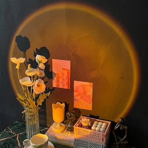 Peut inclure: Un vase en verre cannelé transparent avec des fleurs de pavot blanches, une lampe jaune en forme de tulipe, un sablier, une caisse en plastique rose contenant de petites sphères blanches et une figurine abstraite sombre, sous une lumière de coucher de soleil.