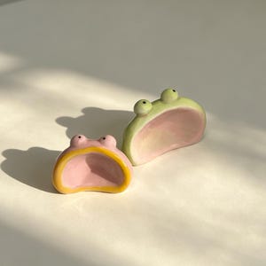 May include: Two ceramic frog-shaped holders. The smaller one is pink with a yellow rim, and the larger one is light green with a pink interior. Both feature two small, raised eyes on top and a wide, open mouth design for holding small items.