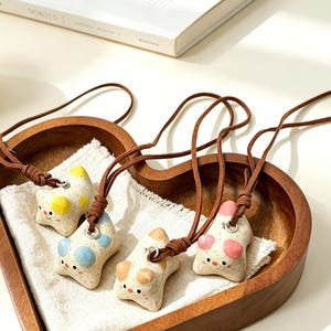 May include: Four star-shaped pendants made of speckled off-white material, each featuring a unique pastel-colored accent on the top points and a simple face with two small eyes and a mouth. Each pendant hangs from a brown cord necklace.