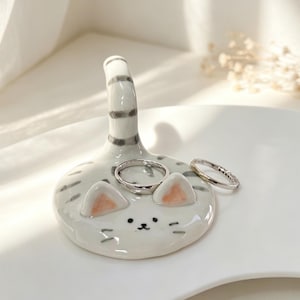 May include: A ceramic ring holder in the shape of a cat, with a tail and a face. The cat has a white base with grey stripes and small, peach-colored ears. Two rings are placed on the holder.