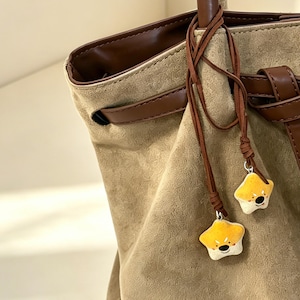May include: A tan suede-textured tote bag featuring dark brown leather-like trim, a decorative belt detail, and two thin brown cords hanging from the handle, each adorned with a small, star-shaped charm depicting a smiling cartoon bear face.