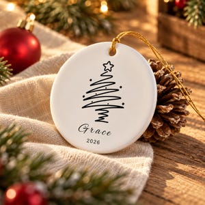Peut inclure: Une décoration de Noël ronde en céramique blanche ornée d'un sapin de Noël dessiné en lignes noires avec une étoile au sommet. Le prénom Grace et l'année 2026 sont inscrits en noir. Un cordon doré est fixé au sommet pour la suspension.