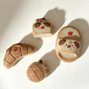 May include: Four ceramic pieces shaped like baked goods with dog faces. Included are a croissant, a round bun, a square loaf, and an oval piece, all featuring speckled tan surfaces, brown ears, and small red noses painted on the front.