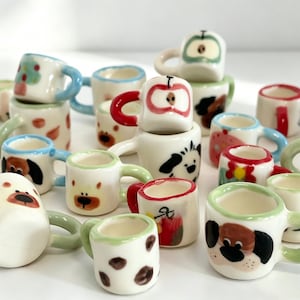 May include: A collection of miniature ceramic mugs featuring various hand-painted designs including animal faces, fruits, and abstract patterns. Each small cup has a rounded handle and a smooth, glossy finish, displayed in a group on a white surface.
